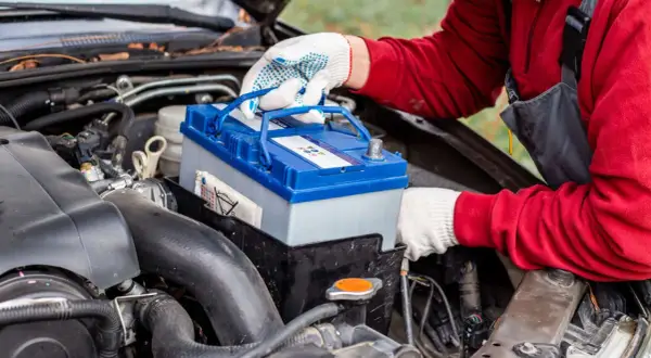 Persona con guantes blancos manipula una batería de coche azul y gris, lista para cargar batería coche.
