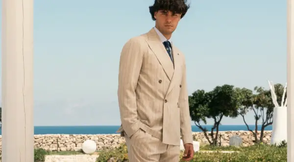 Uomo elegante con abito gessato beige, in piedi con lo sguardo rivolto verso l'osservatore, con il mare e il cielo azzurro sullo sfondo.