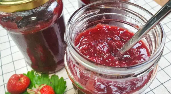 Słoiki z domowym dżemem truskawkowym, łyżeczka w jednym słoiku, świeże truskawki i liście.