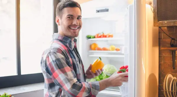 Hombre sonriente saca pimientos de la nevera. Se pregunta cuánto dura el pisto en la nevera, mientras organiza sus verduras.