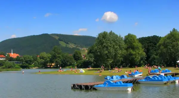 Słoneczny dzień nad jeziorem w Międzybrodziu Żywieckim. Ludzie odpoczywają na plaży, a przy brzegu cumują niebieskie rowerki wodne.