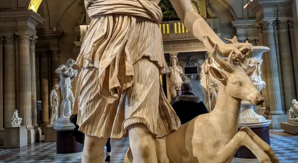 Statue de Diane chasseresse, qui est Artémis, tenant un cerf. L'œuvre est dans un musée, avec d'autres sculptures en arrière-plan.