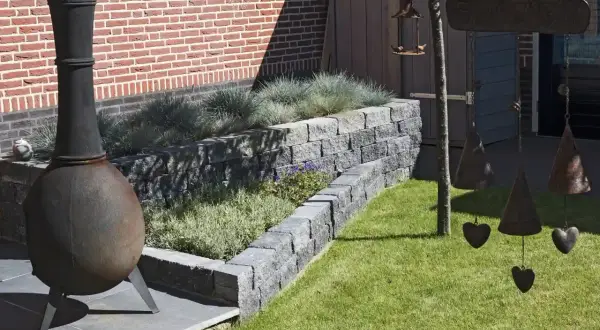 Ein Garten mit einem rustikalen Ofen und einer schönen Mauer aus **Pflanzsteinen setzen** für die Bepflanzung. Dekorative Glocken hängen von einem Baum.