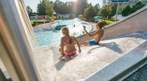 Kinder rutschen im Freibad Leutkirch auf einer Wasserrutsche. Im Hintergrund das Schwimmbecken mit Badegästen.