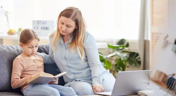 Kobieta i dziecko siedzą na kanapie, dziecko czyta książkę, obok leży laptop.