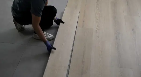 Persona con guantes instala piso SPC de madera clara sobre suelo gris.
