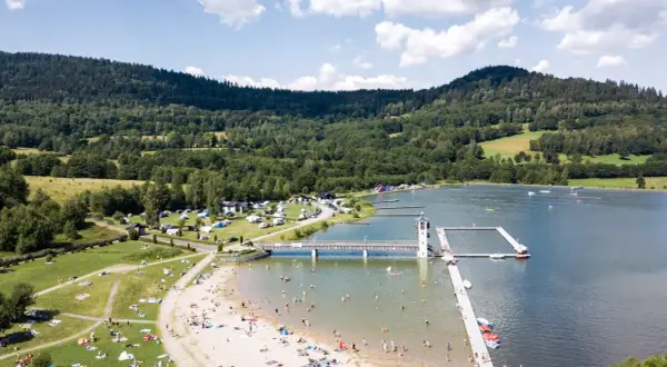 Słoneczny dzień nad zalewem Stara Morawa. Ludzie odpoczywają na plaży i w wodzie, a w tle widać las i pola namiotowe.