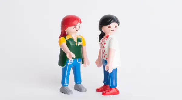 Twee Playmobil-poppetjes staan tegenover elkaar. De ene met rood haar en een groen vest, de ander met zwart haar en een wit shirt. Ze lijken te praten, alsof ze elkaar vertellen dat ik hoor niet bij mijn familie.