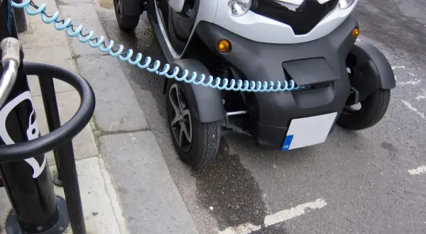 Biały, trójkołowy pojazd elektryczny podłączony do ładowarki. To pokazuje, jak zbudować samochód elektryczny, zaczynając od ładowania.