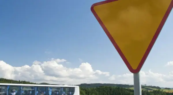 Żółty trójkątny znak a7 ostrzega przed ustąpieniem pierwszeństwa. W tle jedzie autobus, a za nim zielone wzgórza.
