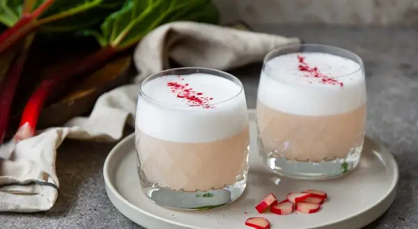 Två glas med krämig rabarber sockerlag, toppade med skum och strössel. Färska rabarberstjälkar i bakgrunden.