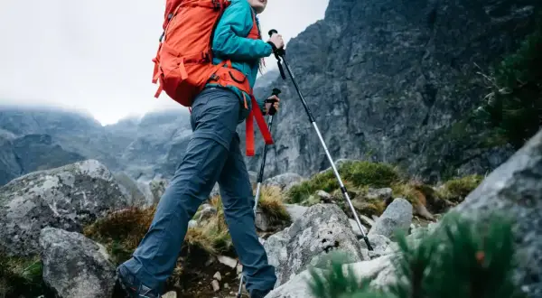 Kobieta z plecakiem wyrusza na wycieczkę w góry. Używa kijków trekkingowych, by pokonać kamienisty szlak.