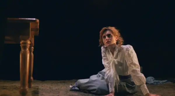 La signorina Julie, con capelli rossi e un vestito bianco, è inginocchiata su un pavimento di terra, con uno sguardo perso nel vuoto.