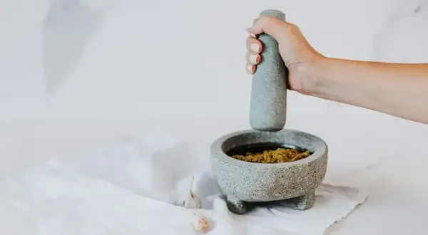 Dłoń uciera składniki w moździerzu, pokazując, jak zrobić pesto. Obok leżą ząbki czosnku.