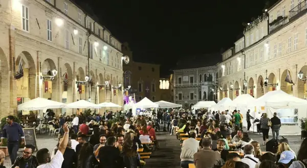 Serata di festa in una piazza delle Marche, con tavoli all'aperto e gente che mangia e socializza.