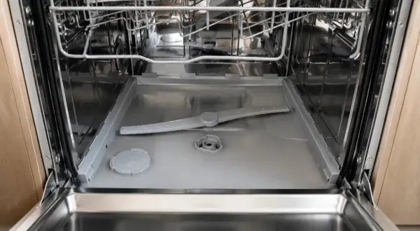 Intérieur d'un lave-vaisselle ouvert, montrant le bras d'aspersion et le fond. On observe de l'eau stagnante au niveau du filtre, signe d'un nettoyage nécessaire.
