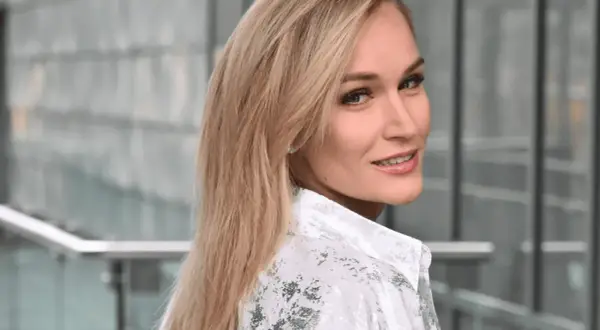 Uśmiechnięta Kamila Boś, w białej koszuli z wzorem, patrzy przez ramię.