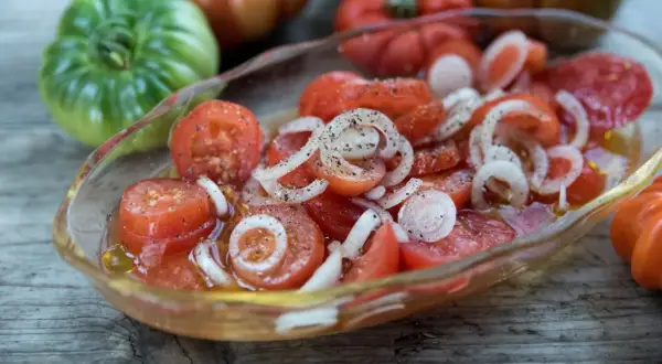 En skål med skivad tigerella tomat och lök, kryddad med peppar. Bakom syns fler tomater i olika färger.