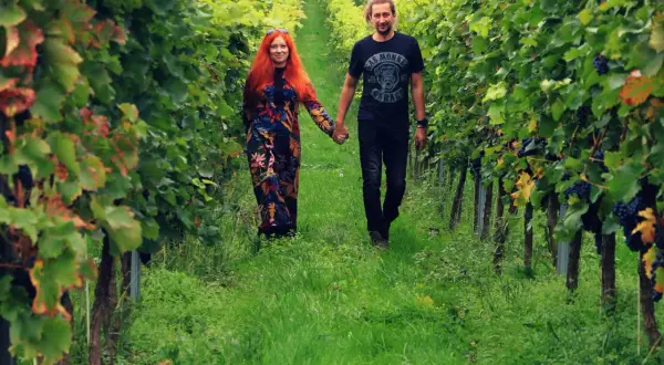 Para spaceruje po winnicy na Dolnym Śląsku, trzymając się za ręce. Wokół bujna zieleń i dojrzewające winogrona.