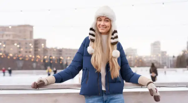 Uśmiechnięta blondynka w granatowej kurtce i białej, futrzanej czapce z pomponami. Idealny zestaw na lodowisko.