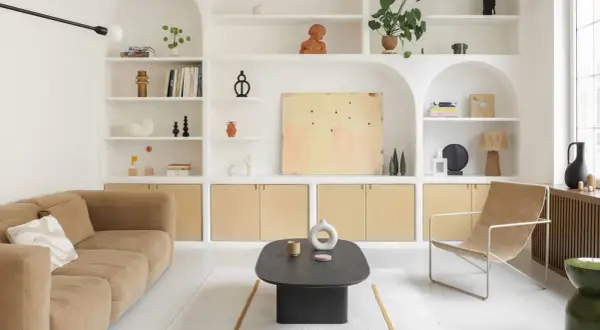 Salon épuré avec canapé beige, fauteuil en toile, table basse noire et étagères murales remplies d'objets décoratifs. Le tout baigne dans une douce lumière, rehaussée par la **couleur blanc cassé** des murs et des étagères.