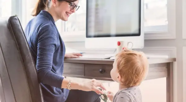 En kvinna med glasögon ler mot sitt barn vid ett skrivbord. Hon visar vad betyder flexibel genom att balansera arbete och familjeliv.