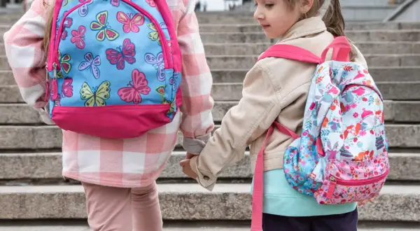 Dwie dziewczynki w kolorowych plecakach szkolnych trzymają się za ręce na schodach.