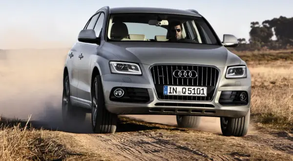 Srebrny Audi Q5 z kierowcą w okularach jedzie po piaszczystej drodze, wzbijając kurz. Wiele osób szuka audi q5 opinie przed zakupem.