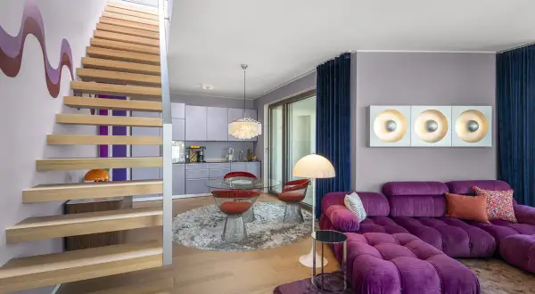 Ein Wohnbereich im 70er jahre einrichtungsstil mit lila Sofa, runden roten Stühlen und einer Holztreppe.