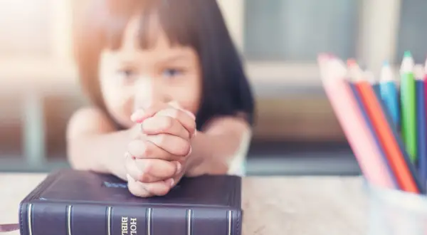 Dziecko z modlącymi się dłońmi na Biblii, obok kolorowe kredki. Nauka 10 przykazań bożych dla dzieci.