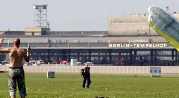 Ein Mann mit nacktem Oberkörper steht auf dem Feld vor dem BERLIN-TEMPELHOF. Ein Gleitschirm liegt im Vordergrund.