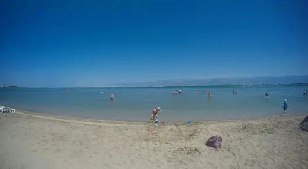 Piaszczysta plaża w Chorwacji z ludźmi kąpiącymi się w morzu i dzieckiem bawiącym się na piasku.