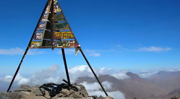 Sommet du mont Toubkal, altitude impressionnante. Le cairn triangulaire, couvert de graffitis, se dresse fièrement sur un pic rocheux, dominant un paysage montagneux spectaculaire sous un ciel bleu éclatant.