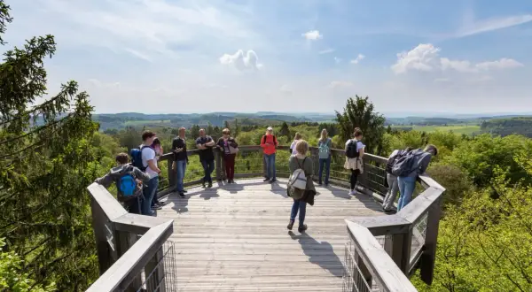 Besucher genießen die Aussicht vom Baumwipfelpfad NRW. Eine Gruppe steht auf der Holzplattform, umgeben von grünen Wäldern und blauen Himmel.