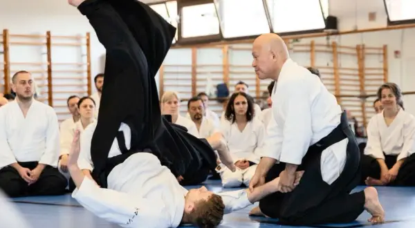 Två personer i vit gi utför en kastteknik inom kampsporten aikido. Åskådare sitter på golvet och observerar.