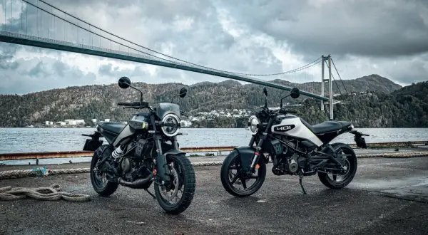 Dwa motocykle Husqvarna Svartpilen 250 stoją nad wodą, pod mostem wiszącym, w malowniczym, górzystym krajobrazie.