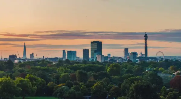 London im Sommer: Die Skyline mit dem Shard und dem BT Tower ragt über einem grünen Park auf, während die Sonne den Himmel in warme Farben taucht.