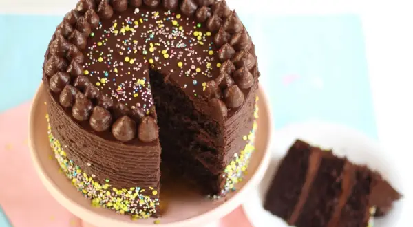 Eine köstliche Schokocreme Torte mit Schokoglasur, Schokotuffs und bunten Streuseln. Ein Stück ist bereits auf einem Teller angerichtet.