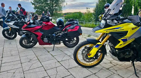 Kilka motocykli Suzuki zaparkowanych na brukowanym placu, z ludźmi w tle.