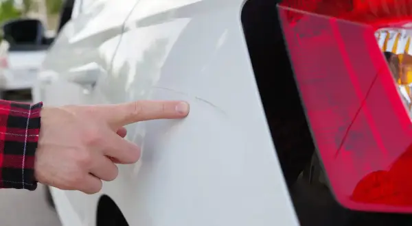 Finger zeigt auf tiefe Kratzer bis auf Grundierung am weißen Auto.