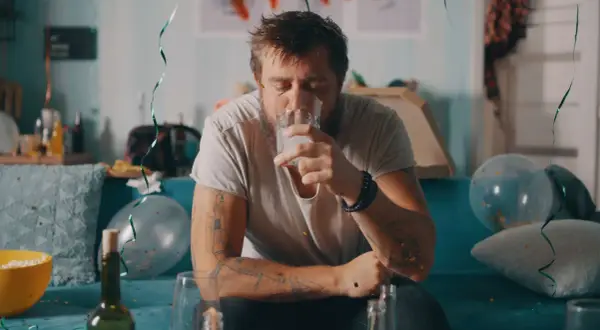 Zmęczony mężczyzna z tatuażami pije wodę po imprezie, symbolizującą jego odstawienie alkoholu. W tle resztki zabawy.