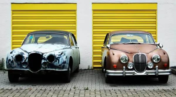 Zwei Oldtimer vor gelben Garagentoren. Einer ist stark verrostet, der andere glänzt. Überlegen Sie, ob auto selber lackieren kosten günstiger sind.