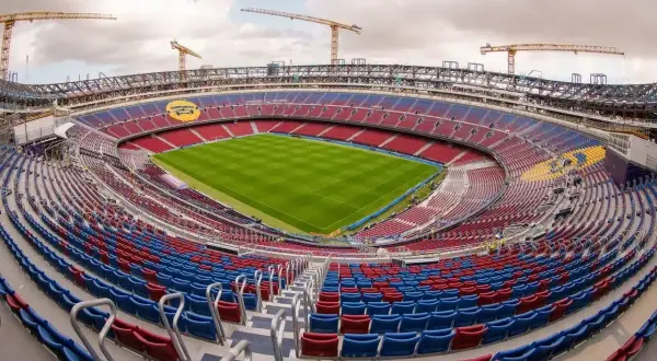 Widok na stadion w budowie, z czerwono-niebieskimi trybunami i zieloną murawą. W tle widoczne są dźwigi, sugerujące, że to jeden z największych stadionów w Polsce.
