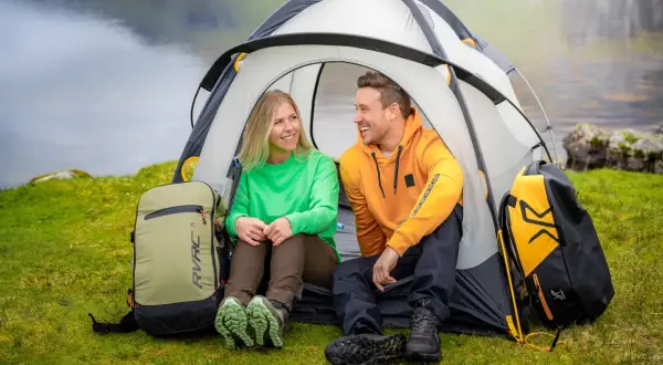 Ett glatt par njuter av att campa i skogen vid en sjö. De sitter i sitt tält med ryggsäckar bredvid.