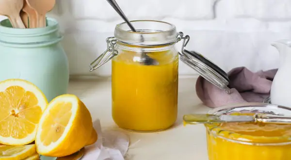 Vasetto di vetro pieno di lemon curd, con limoni freschi affettati e un cucchiaino.