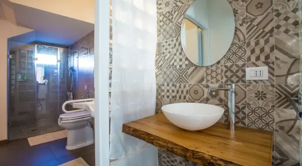 Bagno piccolo con antibagno, doccia in vetro, WC e lavabo su mensola in legno grezzo. Piastrelle geometriche e specchio rotondo.