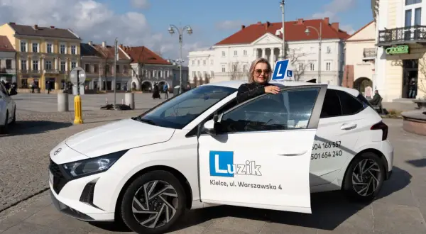 Kobieta z uśmiechem opiera się o drzwi białego samochodu szkoleniowego z literą "L" na dachu. To idealny moment, by zacząć naukę jazdy i zdobyć prawo jazdy B.