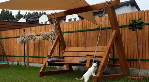 Drewniana huśtawka w ogrodzie, z beżowym daszkiem i poduszkami. Biały kotek przygląda się aranżacji.