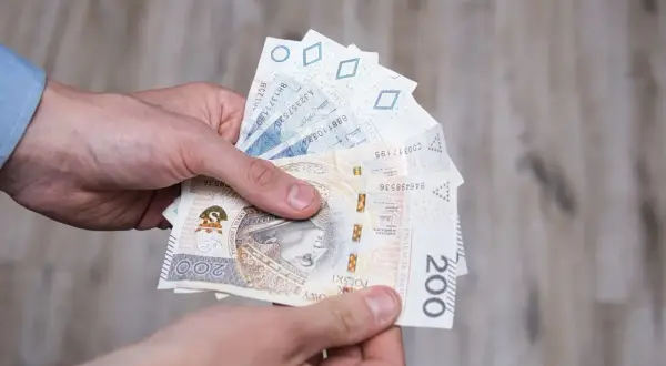 Dłonie trzymają polskie banknoty, w tym 200 zł. Może to symbolizować otrzymanie renty socjalnej z MOPS.