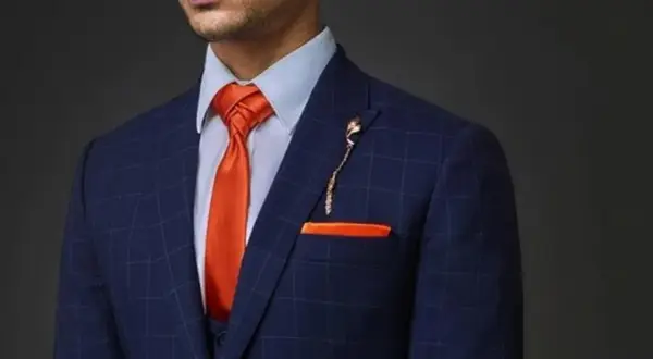 Uomo con abito blu a quadri, camicia azzurra e cravatta arancione con nodi cravatta particolari. Taschino e spilla coordinati.